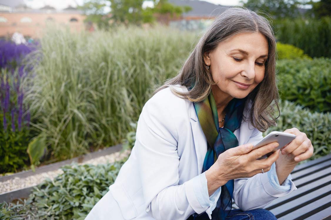 Summertime image of gray haired beautiful retired female sitting on bench with portable electronic gadget, chatting online via messenger. Smiling elegant mature woman using smart phone outdoors