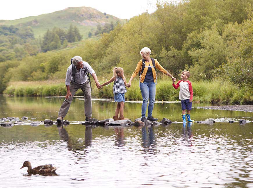Grandparents With Grandchildren Crossing River Whilst Hiking In UK Lake District